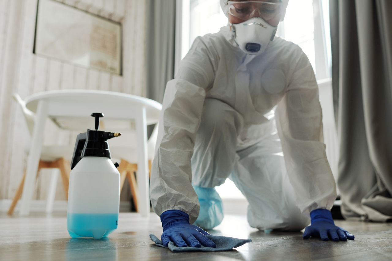 home-hero Person in protective gear cleaning a floor with disinfectant spray and cloth indoors.