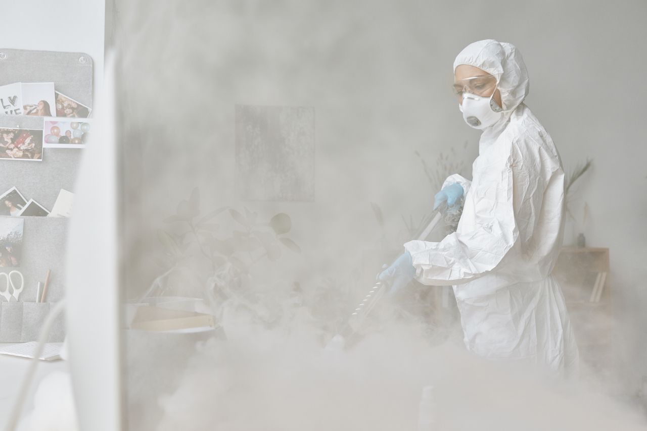 services-05 A woman in a protective suit disinfects an indoor space with fogging equipment.